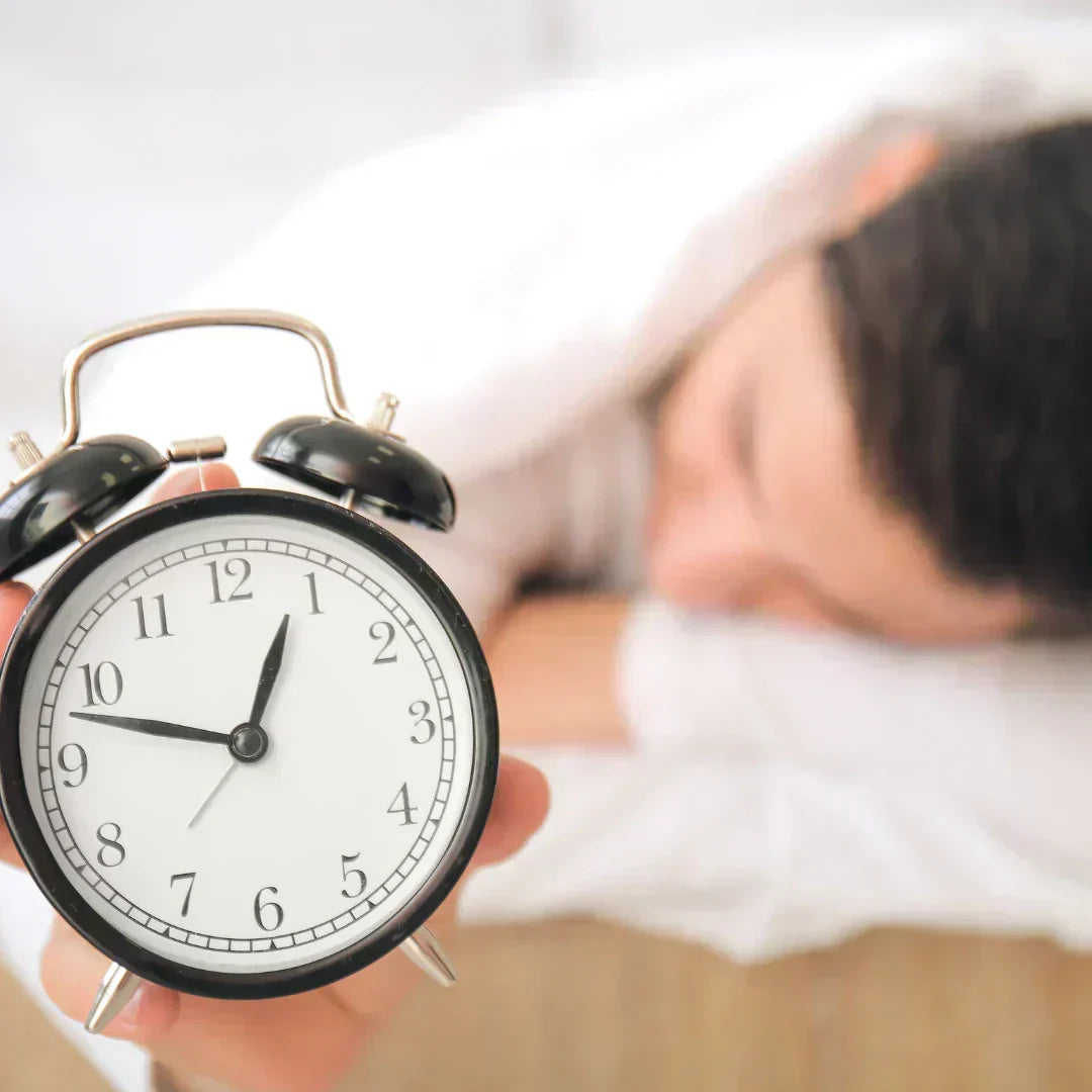 Schwarz-weißer Wecker in Nahaufnahme zeigt 8:50 Uhr; im Hintergrund schläft eine Person – Symbol für geregelte Schlafenszeiten.